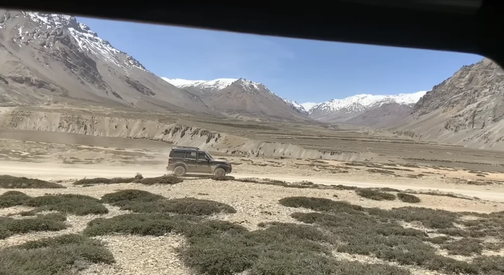 A vehicle driving through a rugged high-altitude valley surrounded by barren mountains and snow-capped Himalayan peaks during the Manali to Ladakh road expedition.