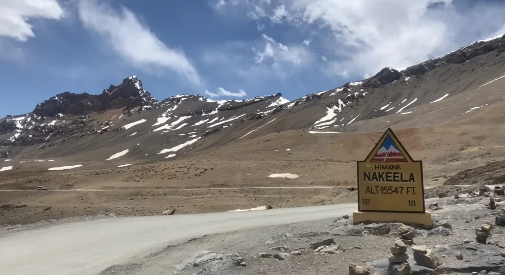 A high-altitude mountain road with the Nakeela Pass sign at 15,547 ft, surrounded by rugged, snow-dusted Himalayan peaks during the Manali to Ladakh road expedition.