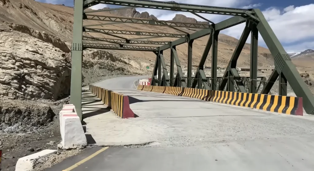 A steel bridge crossing a rugged Himalayan valley, with snow-dusted mountains and winding roads during the Manali to Ladakh road expedition.