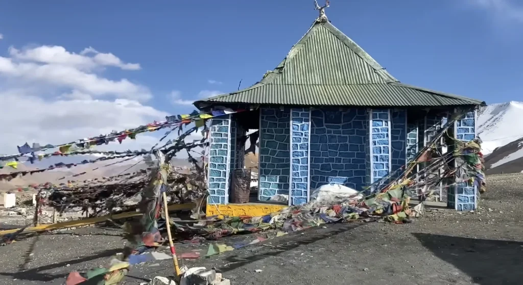 A small blue stone temple decorated with colorful prayer flags, surrounded by snow-covered Himalayan mountains during the Manali to Ladakh road expedition.