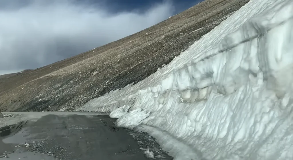 A narrow mountain road bordered by a massive wall of melting snow on a rugged Himalayan slope during the Manali to Ladakh road expedition.