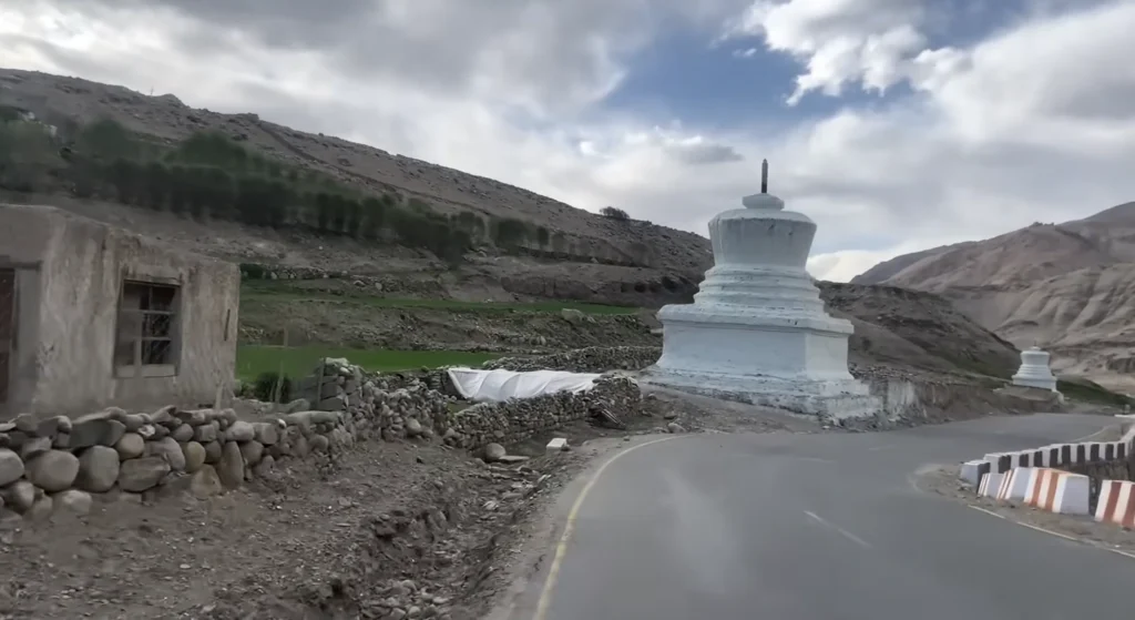 A winding mountain road passing a white Buddhist stupa, with rugged Himalayan terrain, green valleys, and cloudy skies during the Manali to Ladakh road expedition.