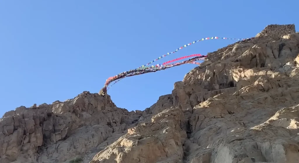 Colorful Buddhist prayer flags stretched across rugged rocky cliffs under a clear blue sky during the Manali to Ladakh road expedition.
