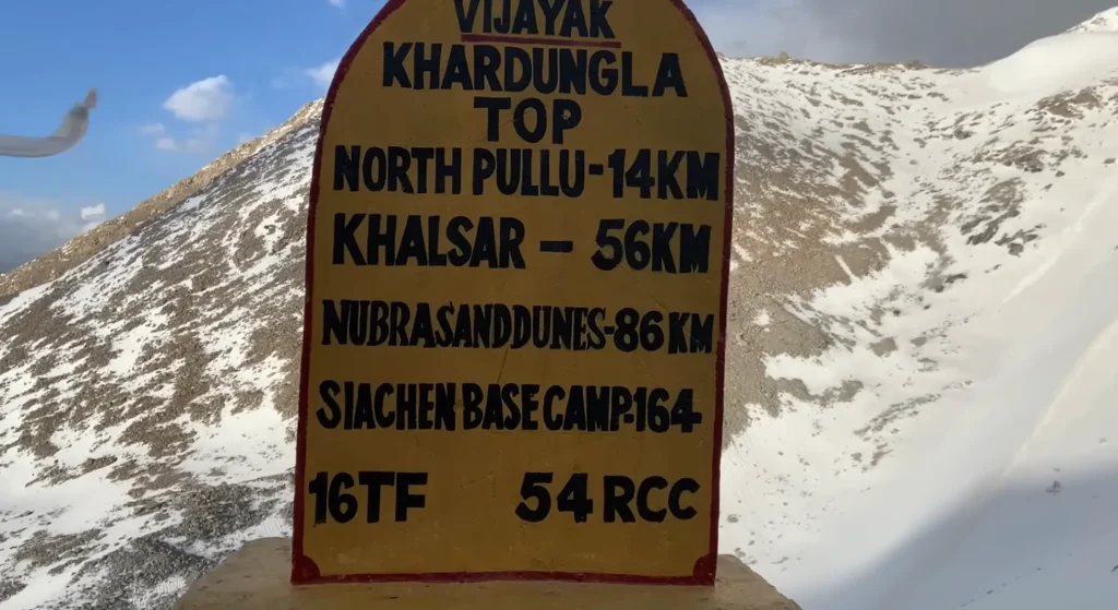 A Khardung La Top sign marking 16,000 ft, surrounded by snow-covered Himalayan mountains during the Manali to Ladakh road expedition.