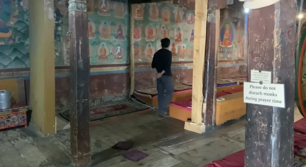 A traditional Buddhist monastery interior with colorful wall paintings, prayer cushions, and a visitor observing the sacred space during the Manali to Ladakh road expedition.