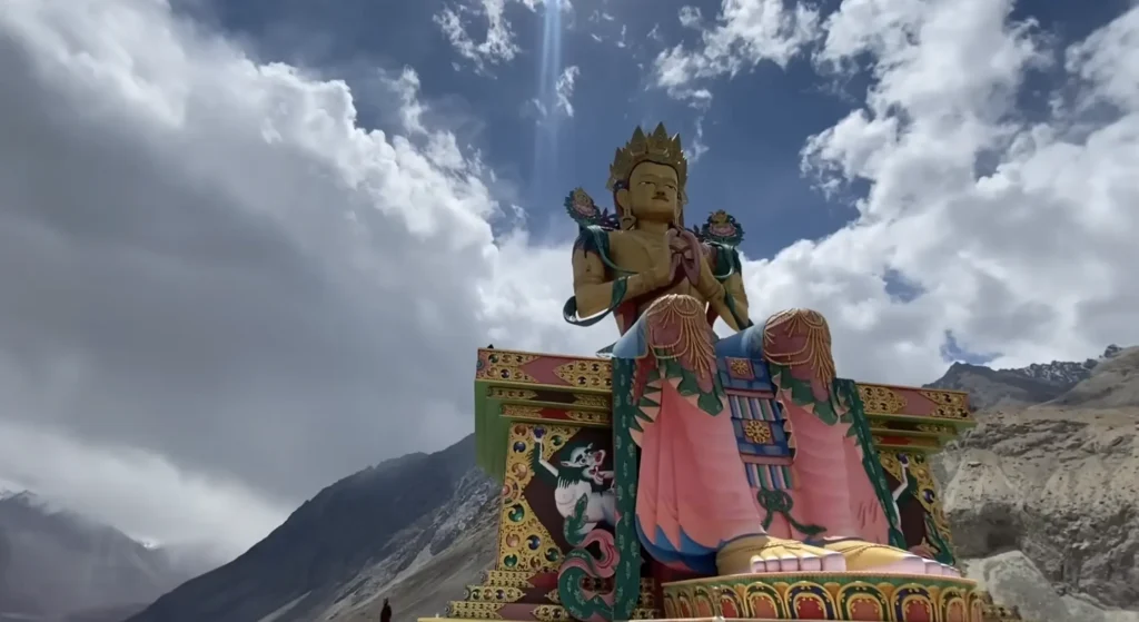 A colorful Buddhist statue set against the dramatic Himalayan mountains and cloudy sky along the Manali to Ladakh road expedition route.