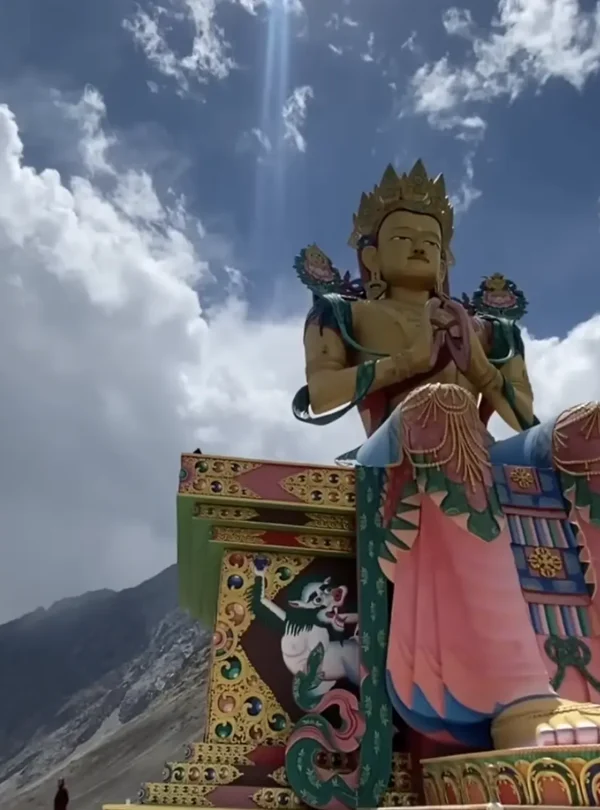 A colorful Buddhist statue set against the dramatic Himalayan mountains and cloudy sky along the Manali to Ladakh road expedition route.