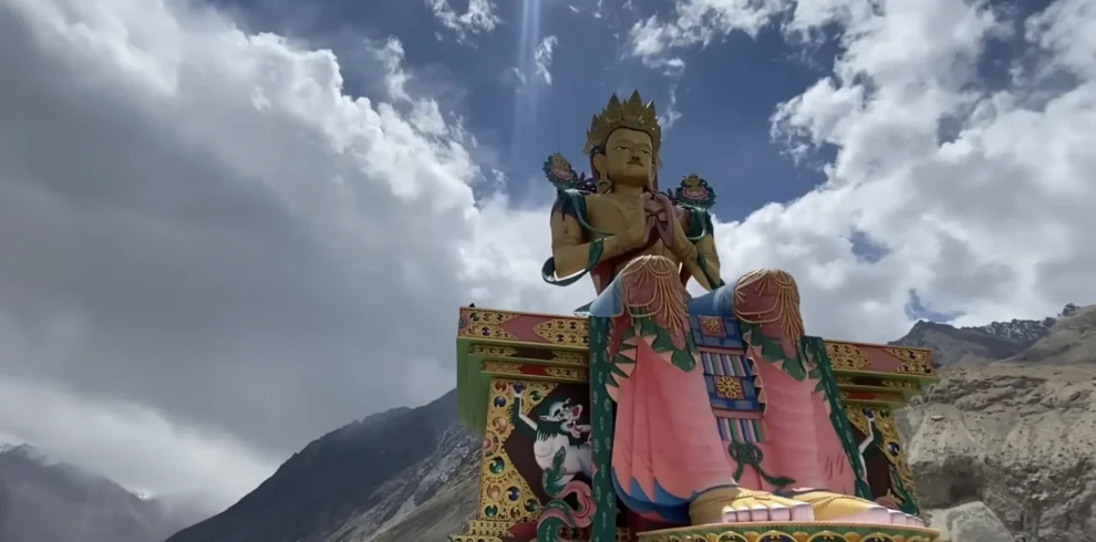 A colorful Buddhist statue set against the dramatic Himalayan mountains and cloudy sky along the Manali to Ladakh road expedition route.