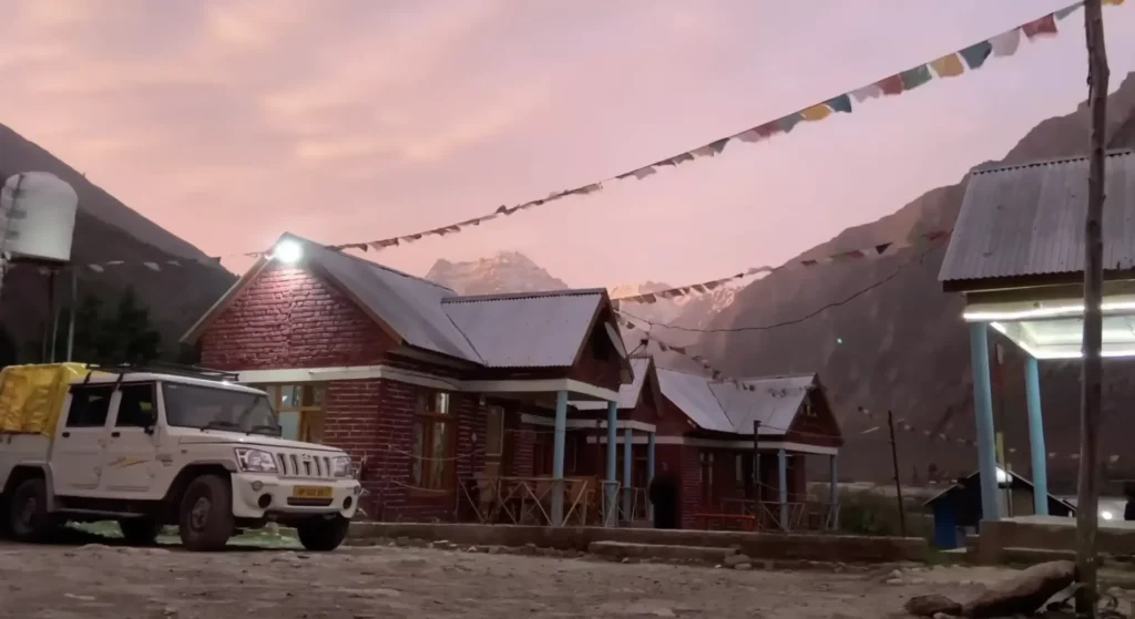 A mountain village with traditional cabins, colorful prayer flags, and a parked vehicle surrounded by the Himalayan landscape at sunset during the Manali to Ladakh road expedition.