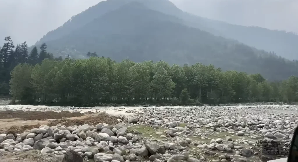 A rocky riverbed with lush green trees and misty Himalayan mountains in the background during the Manali to Ladakh road expedition.
